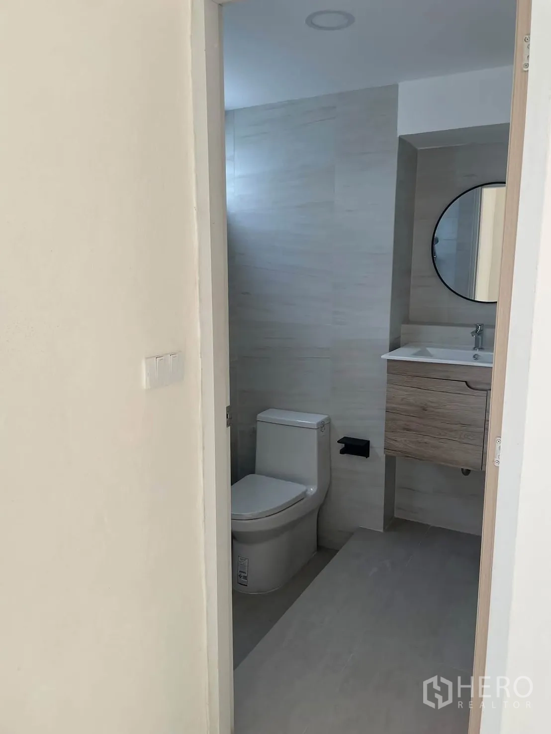 Townhouse for rent in Huai Khwang, Bangkok - Modern bathroom with light tile walls, toilet, vanity cabinet, and round mirror.