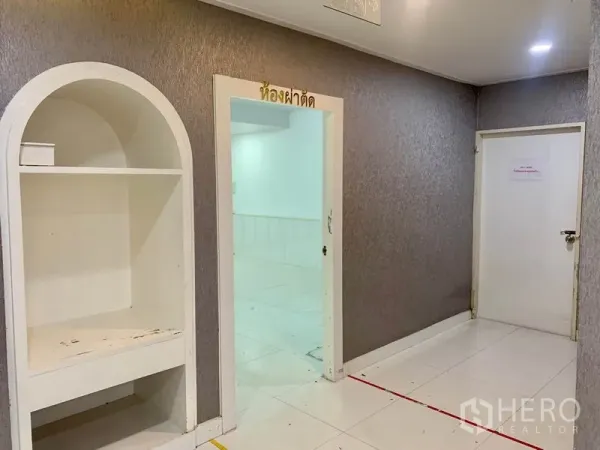 零售空间 出租在Khlong Toei, Bangkok - 室内走廊带拱形嵌入式书架、白色地砖与通往明亮工作区的门。