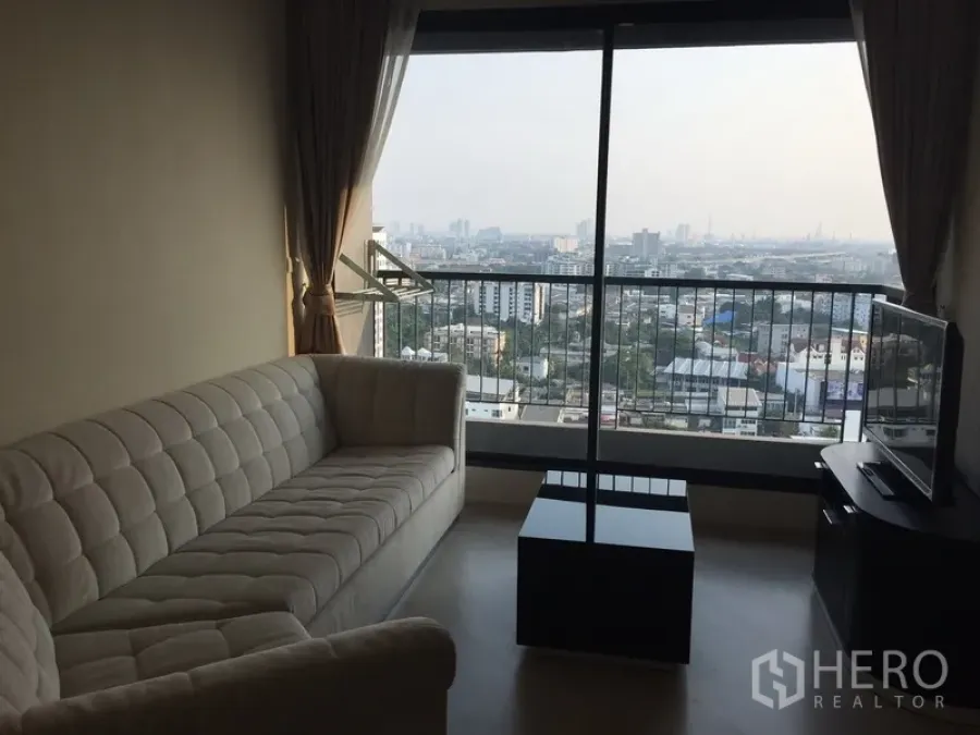 Condominium for rent in Khlong Toei, Bangkok - Bright living room with beige sofa, TV console and floor‑to‑ceiling window offering a wide city view at Rhythm Sukhumvit 44.