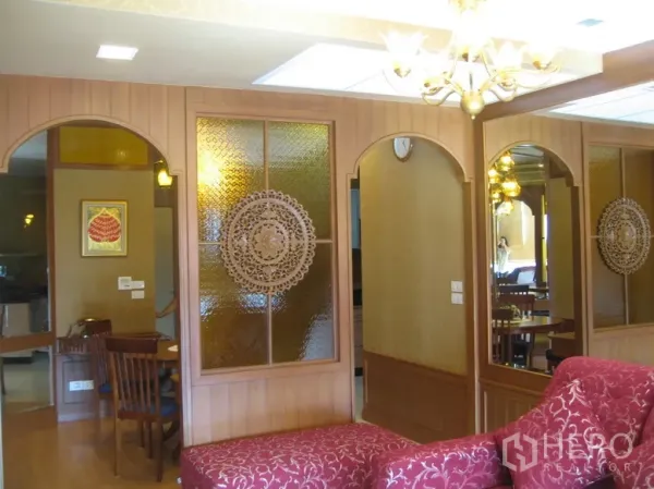 Condominium for rent in Khlong Toei, Bangkok - Living area with warm wood paneling, arched mirrors, decorative frosted glass and a chandelier beside a red chaise.
