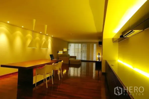 公寓 出租在Watthana, Bangkok - 开放式客餐厅，暖色灯光，长木餐桌、白色椅子，尽头为落地窗与沙发。