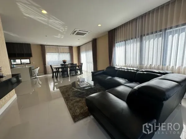 Detached House for rent or sale in Lam Luk Ka, Pathum Thani - Open-plan living and dining area with black leather sectional sofa, dining set and wide windows.