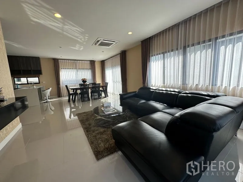 Detached House for rent or sale in Lam Luk Ka, Pathum Thani - Open-plan living and dining area with black leather sectional sofa, dining set and wide windows.