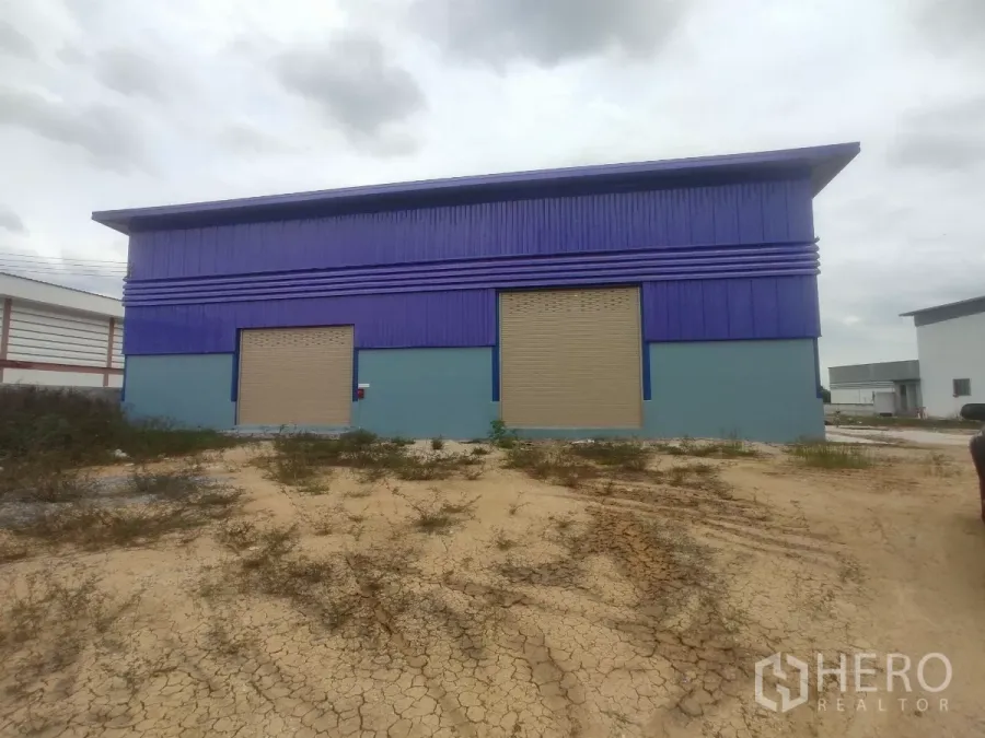 Factory for sale in Khlong Toei, Bangkok - Front view of a blue factory building with two large roller shutter doors and a gravel yard.