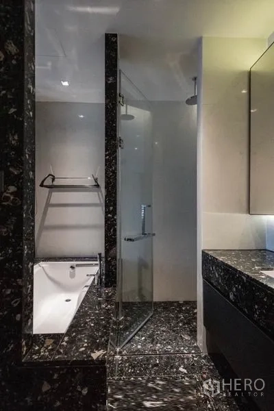 Condominium for rent in Khlong Toei, Bangkok - Bathroom with glass shower, bathtub and dark marble finishes.