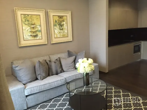 Condominium for rent in Watthana, Bangkok - Open-plan living area with gray sofa, glass coffee table, artwork and modern kitchenette in a Sukhumvit 39 condo.