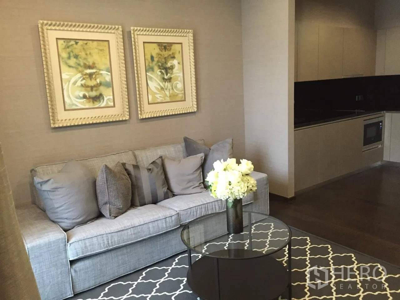 Condominium for rent in Watthana, Bangkok - Open-plan living area with gray sofa, glass coffee table, artwork and modern kitchenette in a Sukhumvit 39 condo.