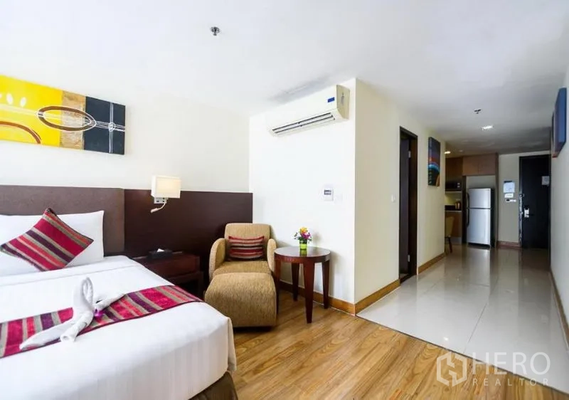 Condominium for rent in Khlong Toei, Bangkok - Bright bedroom with bed, accent pillows, lounge chair and view toward the kitchenette and entry.