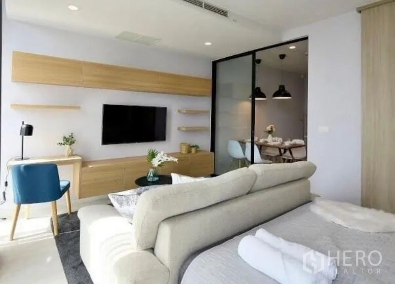 Condominium for rent in Pathum Wan, Bangkok - Living zone with wall-mounted TV, light wood cabinetry, and a sliding glass partition to the dining room.