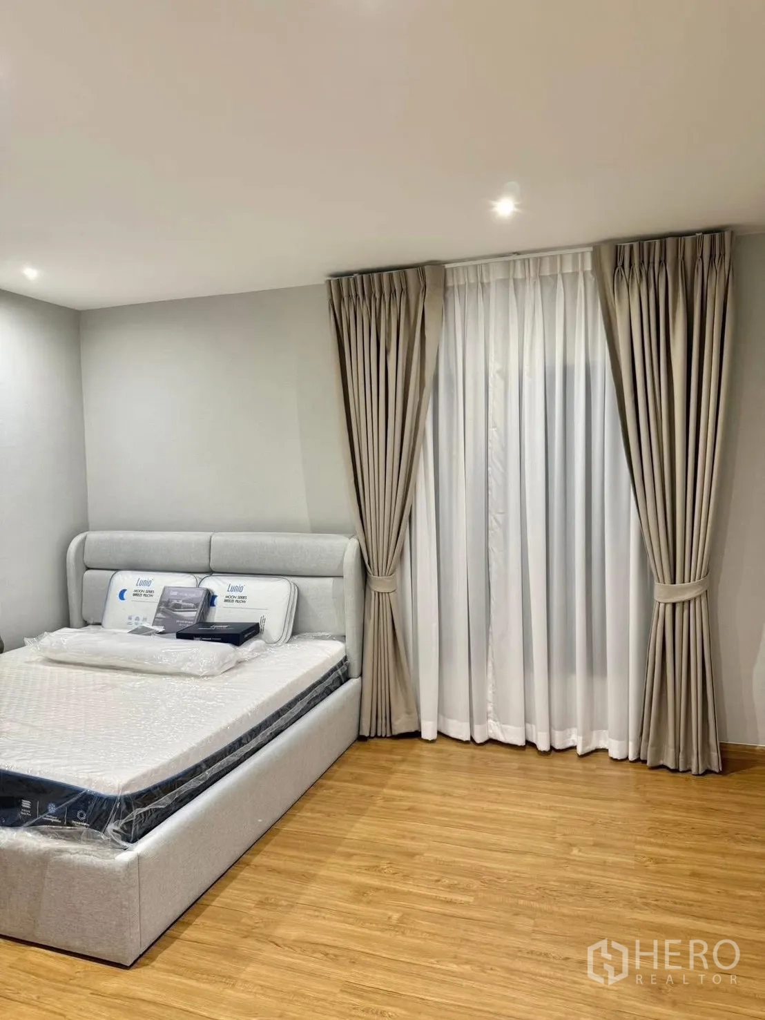 Detached House for rent in Bang Phli, Samut Prakan - Cozy bedroom with grey upholstered bed, floor-to-ceiling curtains and warm wood flooring.