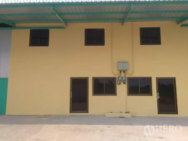工厂 出租在Khlong Luang, Pathum Thani - 工厂正立面：黄色两层办公区，深色窗框，电表箱与遮雨挑檐，前方混凝土硬地