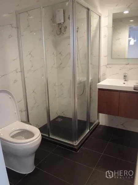 Condominium for rent in Bang Rak, Bangkok - Modern bathroom with a glass-enclosed shower and water heater, vanity with mirror, marble-style walls, and dark floor tiles.