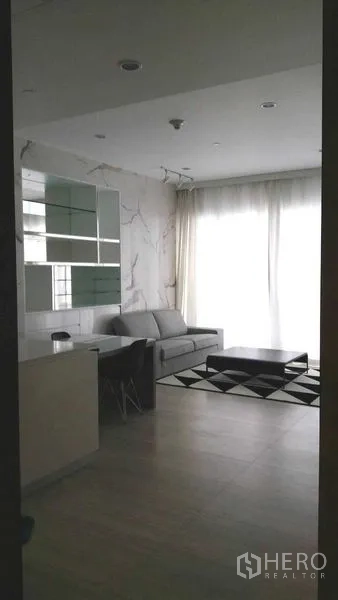 公寓 出租在Pathum Wan, Bangkok - 明亮起居角落，整面窗帘、灰色沙发与黑白地毯上的低矮茶几。