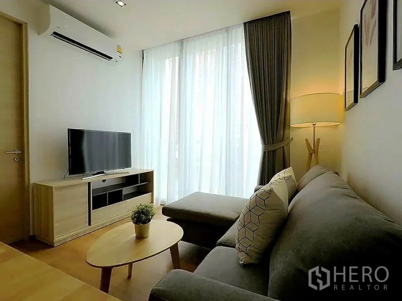 Condominium for rent in Khlong Toei, Bangkok - Compact lounge with L-shaped sofa, TV console and tall sheer curtains.