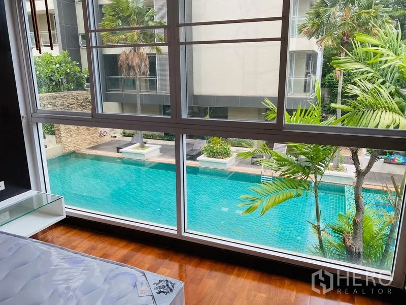 公寓 出租在Watthana, Bangkok - 卧室大窗俯瞰庭院碧蓝泳池与棕榈树景观。