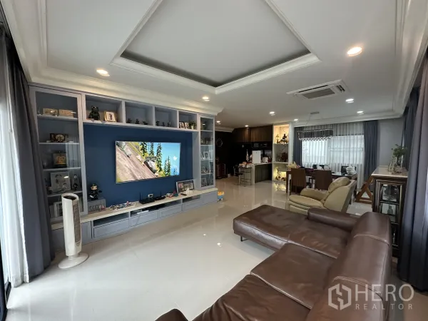 Detached House for rent in Lam Luk Ka, Pathum Thani - Open-plan living room with built-in TV wall and brown leather sectional.
