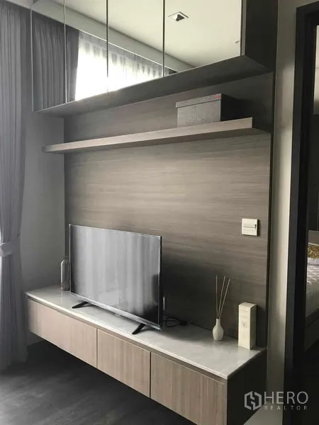 Condominium for rent in Watthana, Bangkok - Living room media wall with flat-screen TV, floating shelf, and built-in cabinets near curtained window.
