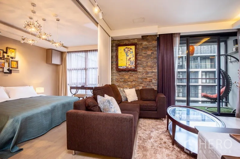 Condominium for rent in Watthana, Bangkok - Living room facing the balcony, L-shaped sofa and glass table, with adjacent loft-style bedroom and feature stone wall.