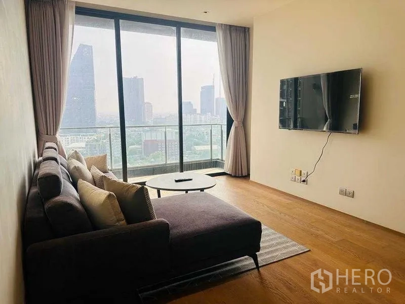 Condominium for rent in Khlong Toei, Bangkok - Bright living area with L-shaped sofa, wall-mounted TV, and balcony skyline view.