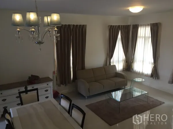 Townhouse for rent in Watthana, Bangkok - Bright living room with beige sofa, glass coffee table, and corner windows with curtains.