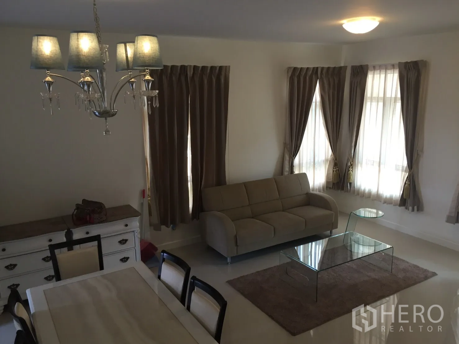 Townhouse for rent in Watthana, Bangkok - Bright living room with beige sofa, glass coffee table, and corner windows with curtains.