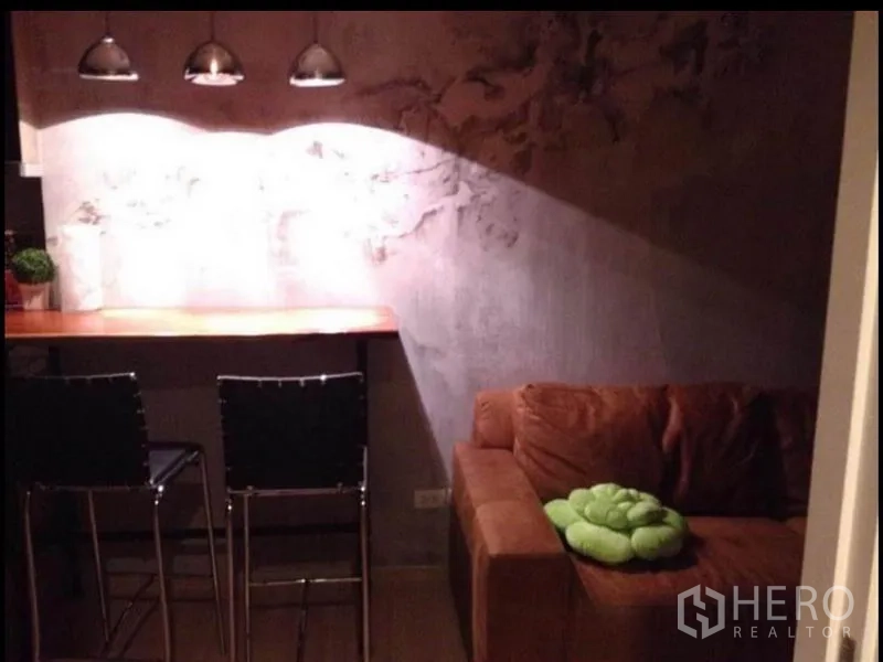 Condominium for sale in Phaya Thai, Bangkok - Compact lounge with brown sofa and breakfast bar illuminated by three pendant lamps.