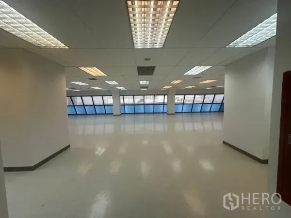 办公空间 出租在Phaya Thai, Bangkok - 开放式办公室，一侧为蓝色斜面带状窗，吊顶荧光灯照明充足。