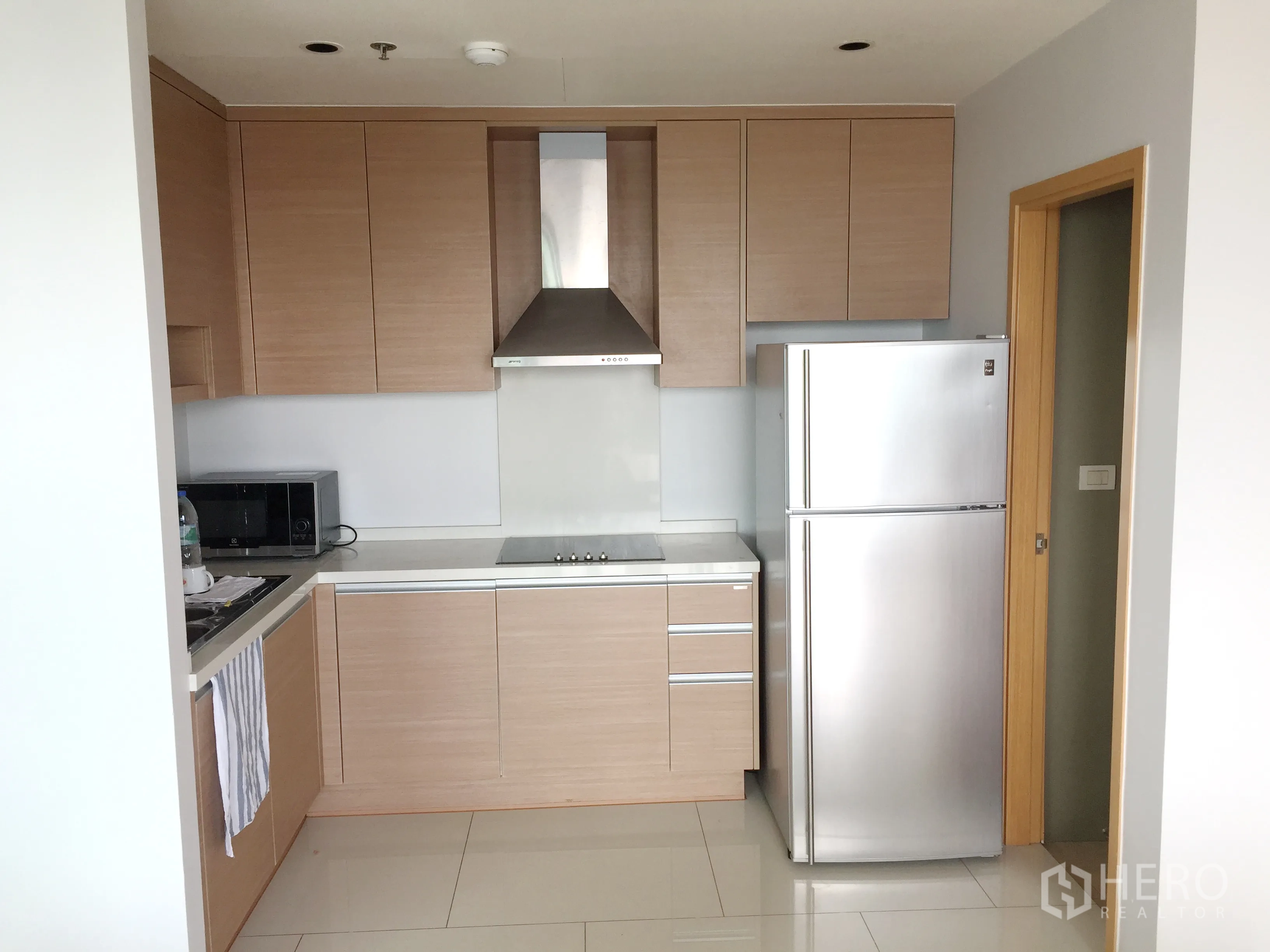 Condominium for rent in Khlong Toei, Bangkok - Modern kitchen with wood cabinetry, cooktop, range hood, microwave, and stainless-steel refrigerator.