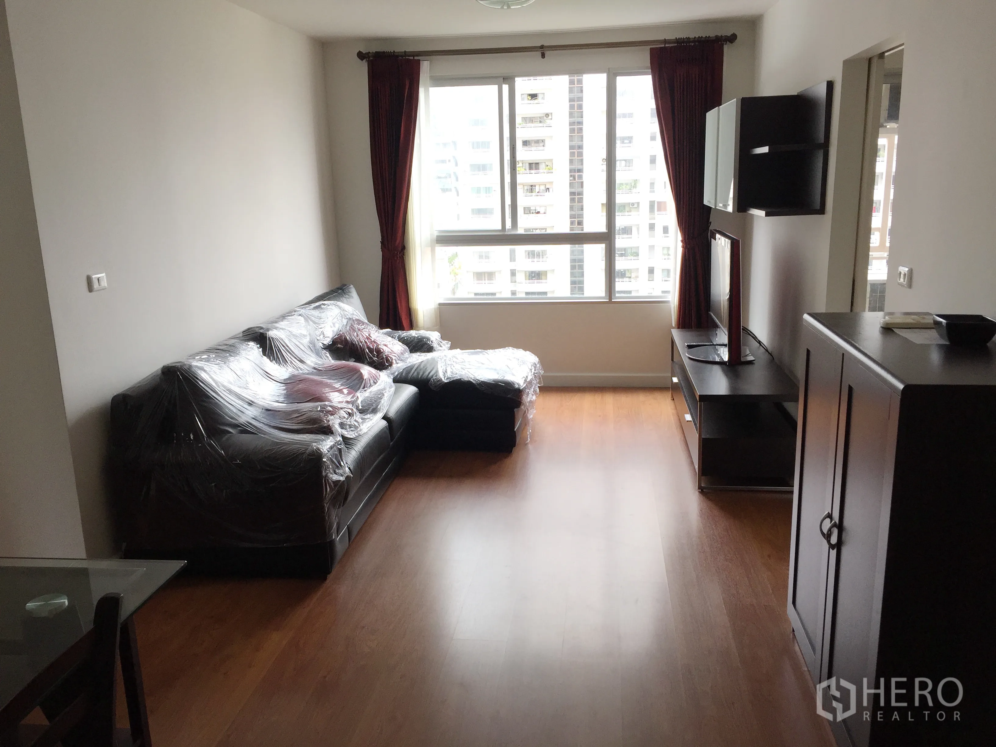 Condominium for rent in Khlong Toei, Bangkok - Bright living room with L-shaped sofa, TV console, wood floors and a large window with city view.