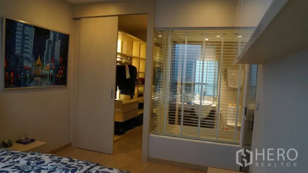Condominium for rent in Watthana, Bangkok - Bedroom looking toward a walk-in closet with sliding door and a glass-partition bathroom in warm wood tones.