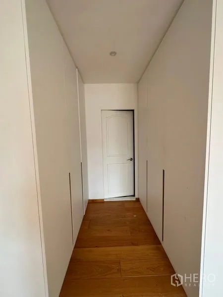 Condominium for rent in Khlong Toei, Bangkok - Hallway with built-in wardrobes on both sides and wooden flooring leading to a white door.