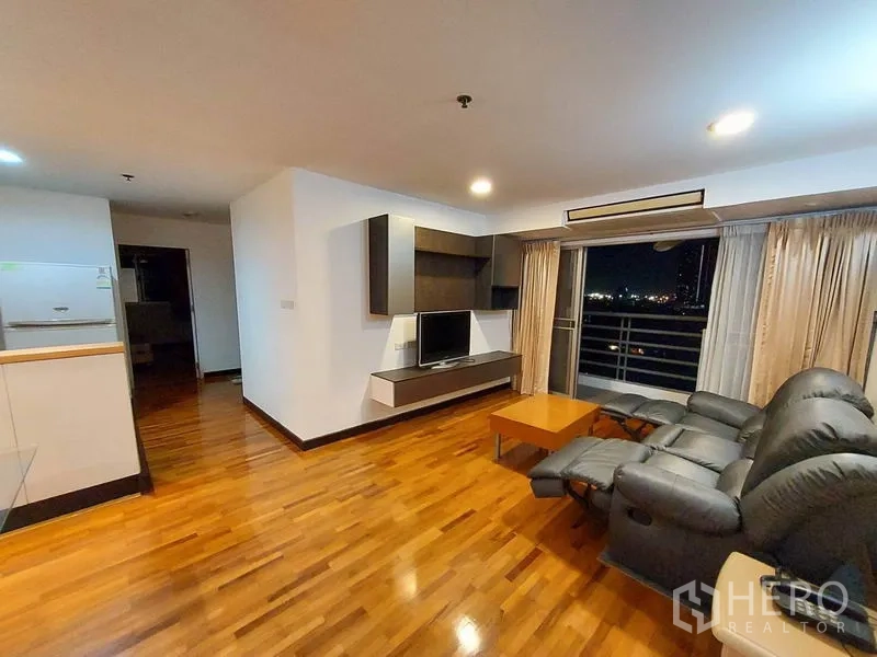 Condominium for rent or sale in Khlong Toei, Bangkok - Living room with reclining sofa, TV console and sliding doors to a balcony with night skyline.