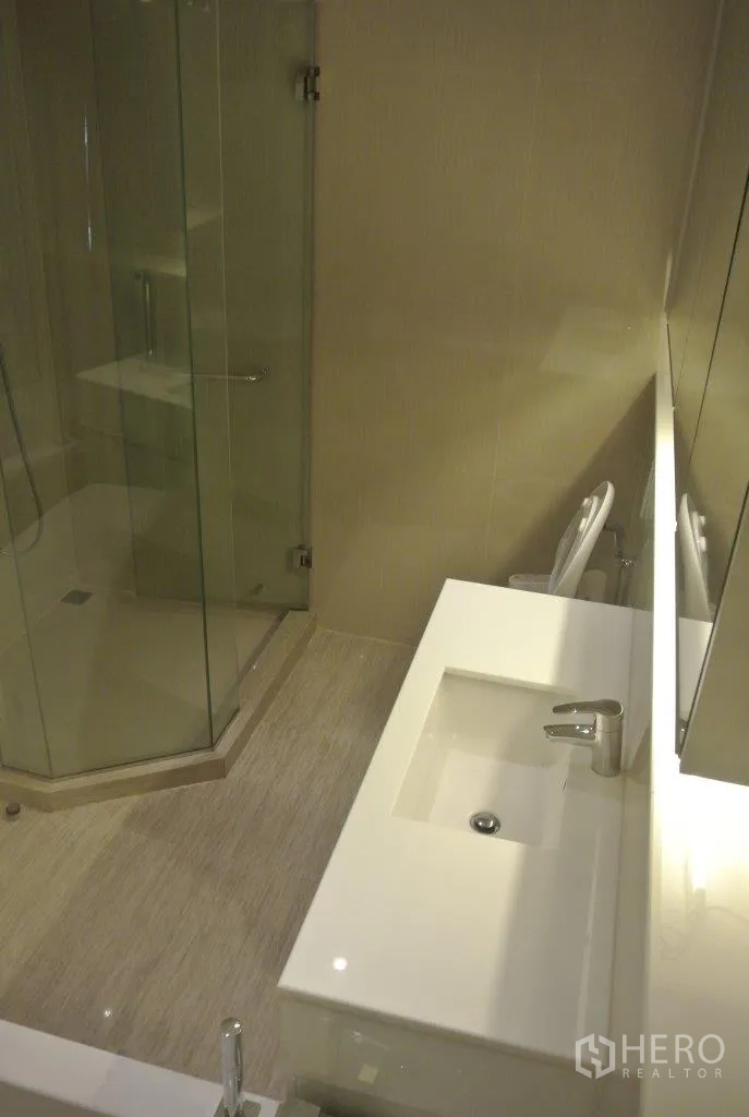 Condominium for rent in Watthana, Bangkok - Bathroom showcasing a glass-enclosed shower, long countertop sink and light beige tiles.