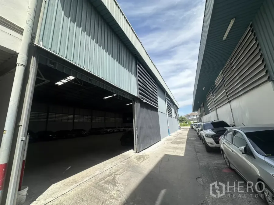 Warehouse for rent in Suan Luang, Bangkok - Wide driveway between metal-clad warehouses with a large sliding door open and cars parked along the side under a blue sky.