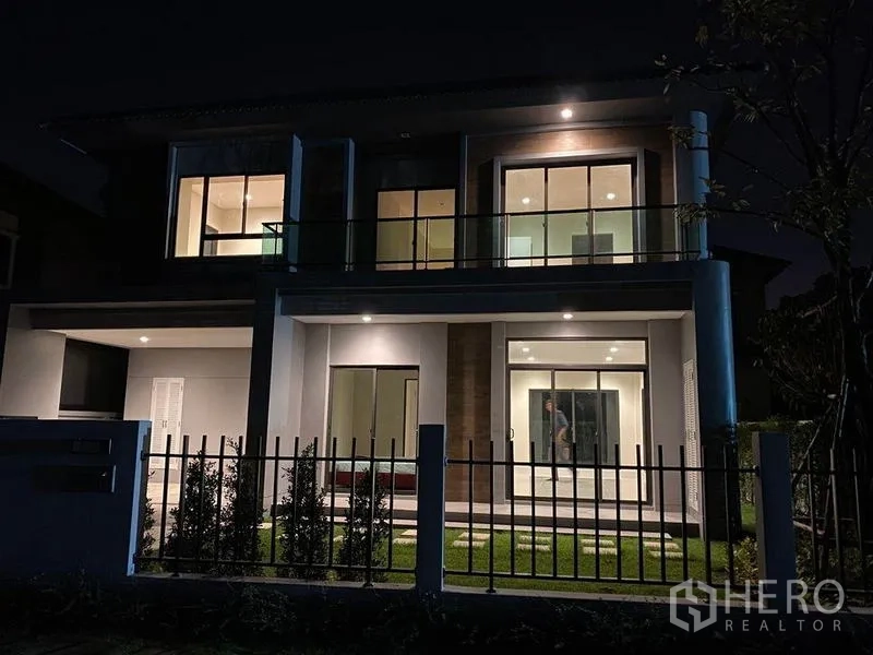 独立式住宅 出租 - 夜间外观，暖色照明、阳台和带围栏的小花园。