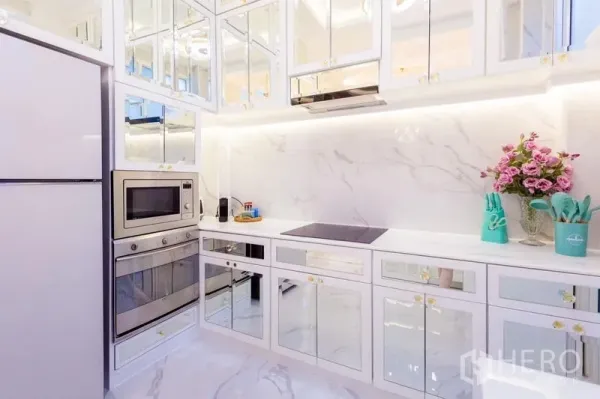 Townhouse for rent in Lat Phrao, Bangkok - Modern white marble kitchen with mirrored cabinets, built-in oven, microwave and induction hob with under-cabinet lighting.