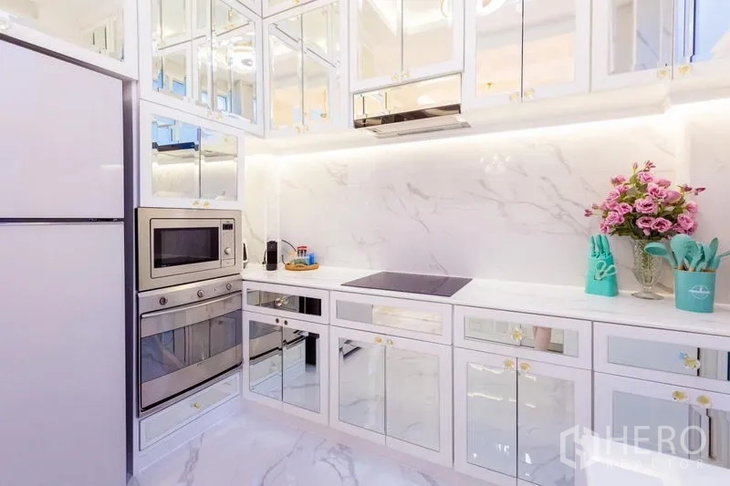 Townhouse for rent in Lat Phrao, Bangkok - Modern white marble kitchen with mirrored cabinets, built-in oven, microwave and induction hob with under-cabinet lighting.