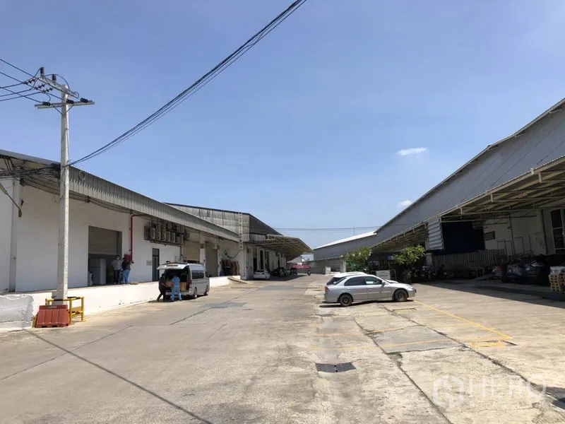 货栈 出租在Mueang Samut Prakan, Samut Prakan - 仓库外部车道，左右为带顶装卸月台的厂房，并设有停车位与电线杆，天空晴朗。