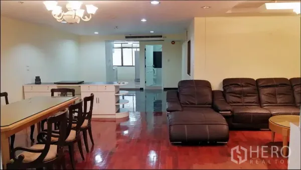 Condominium for rent or sale in Khlong Toei, Bangkok - Open-plan lounge with chandelier lighting, L-shaped sofa, and polished wooden floors.