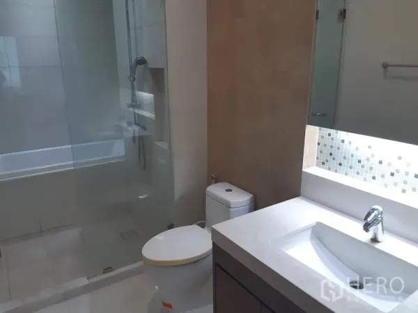 Condominium for rent in Khlong Toei, Bangkok - Modern bathroom with wide vanity, toilet and glass-enclosed shower over bathtub in a Bangkok condo.