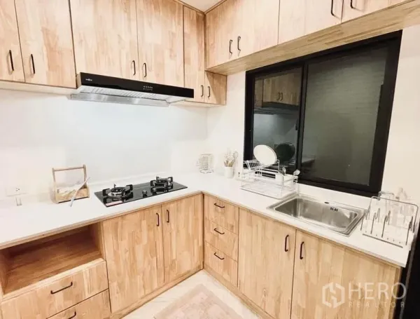 Townhouse for rent in Bang Kapi, Bangkok - L-shaped kitchen with light wood cabinetry, gas hob, range hood, and a stainless-steel sink by a window.