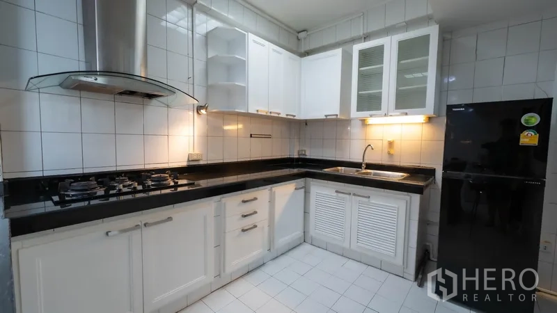Townhouse for rent in Watthana, Bangkok - L‑shaped kitchen with white cabinets, gas hob, hood, and black countertop.