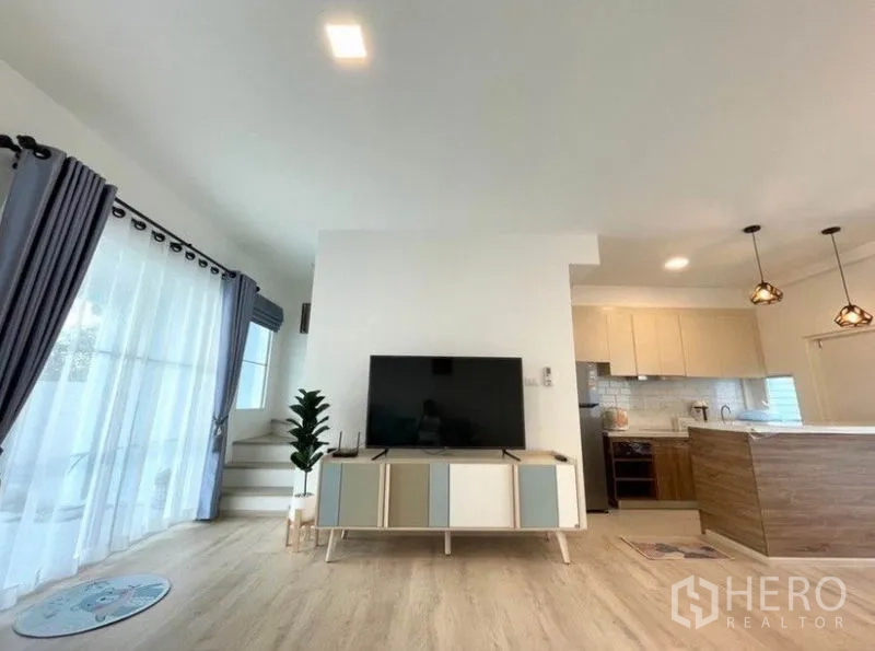联排别墅 for rent in Prawet, Bangkok - 电视柜面向纱帘大窗，旁有楼梯与厨房。