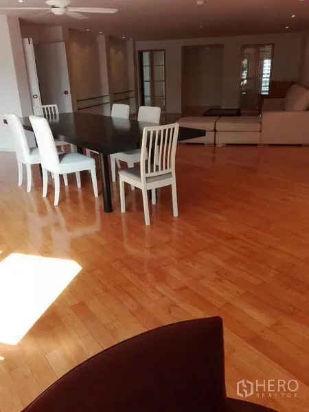Condominium for rent in Khlong Toei, Bangkok - Dining area featuring a long table and white chairs in a large open living space with a ceiling fan and wood floors.