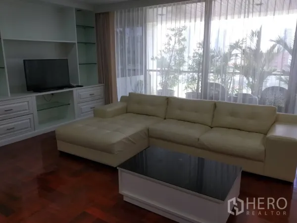 คอนโด ให้เช่าในPathum Wan, Bangkok - ห้องนั่งเล่นโซฟาเข้ามุมสีครีม ตู้ทีวีบิลต์อิน และระเบียงมีต้นไม้หลังผ้าม่านโปร่ง