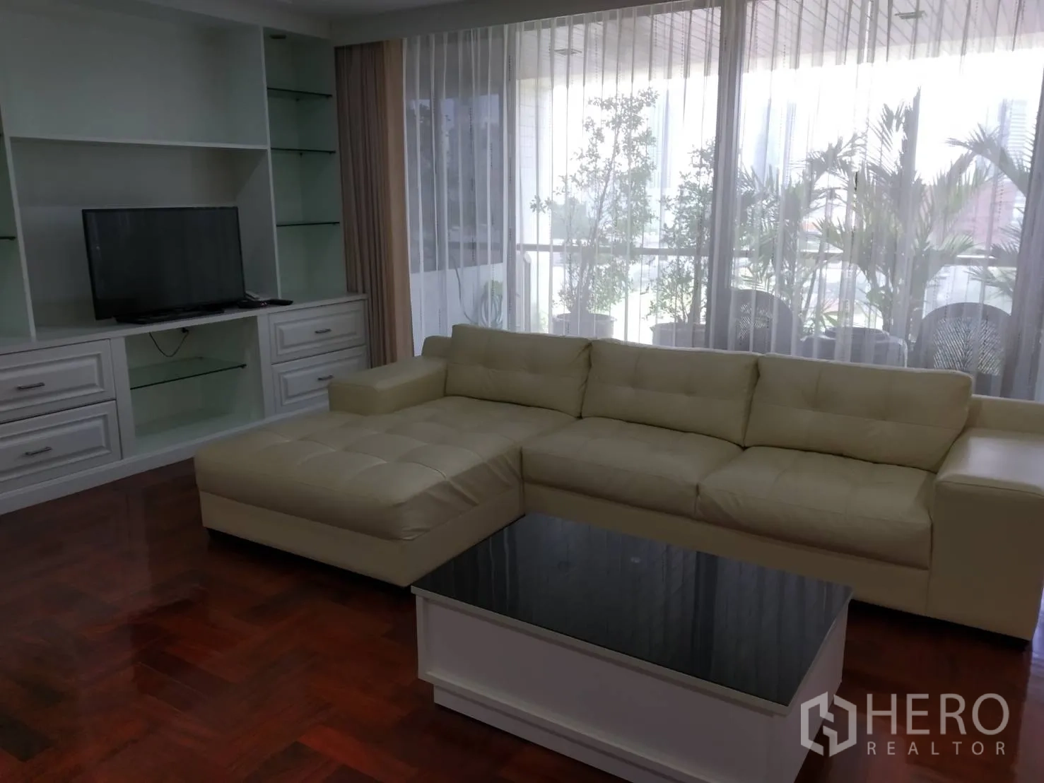 คอนโด ให้เช่าในPathum Wan, Bangkok - ห้องนั่งเล่นโซฟาเข้ามุมสีครีม ตู้ทีวีบิลต์อิน และระเบียงมีต้นไม้หลังผ้าม่านโปร่ง