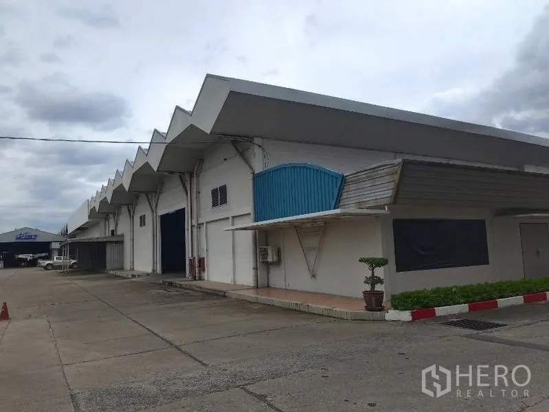Warehouse for rent in Lam Luk Ka, Pathum Thani - Large sawtooth‑roof warehouse exterior with multiple loading doors and wide concrete driveway.