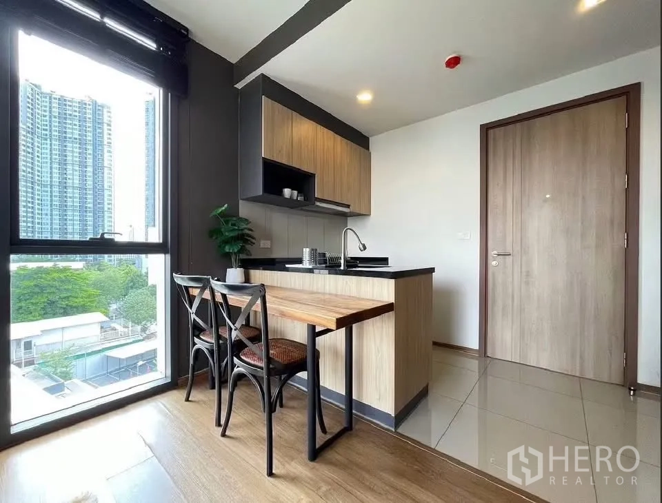 Condominium for rent in Watthana, Bangkok - Open-plan kitchen with wood cabinets, dining table, and a large window with city views.