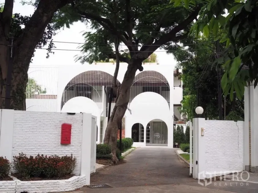 Detached House for rent in Huai Khwang, Bangkok - Gated driveway leading to a white arched building shaded by mature trees.