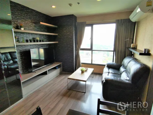 Condominium for rent or sale in Watthana, Bangkok - Living room with black leather sofa, TV console, floating shelves and a large city-view window.